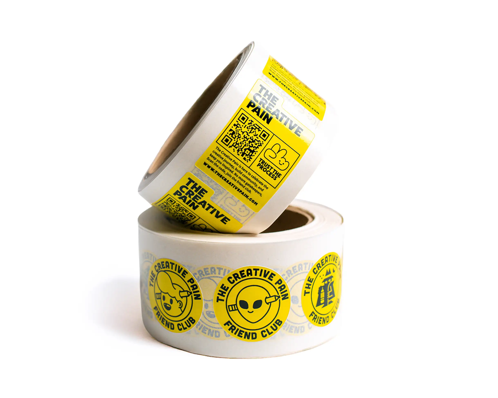 Two rolls of tape with yellow labels. Top roll has text and a QR code, bottom roll features a smiling face and "The Creative Pain Friend Club."