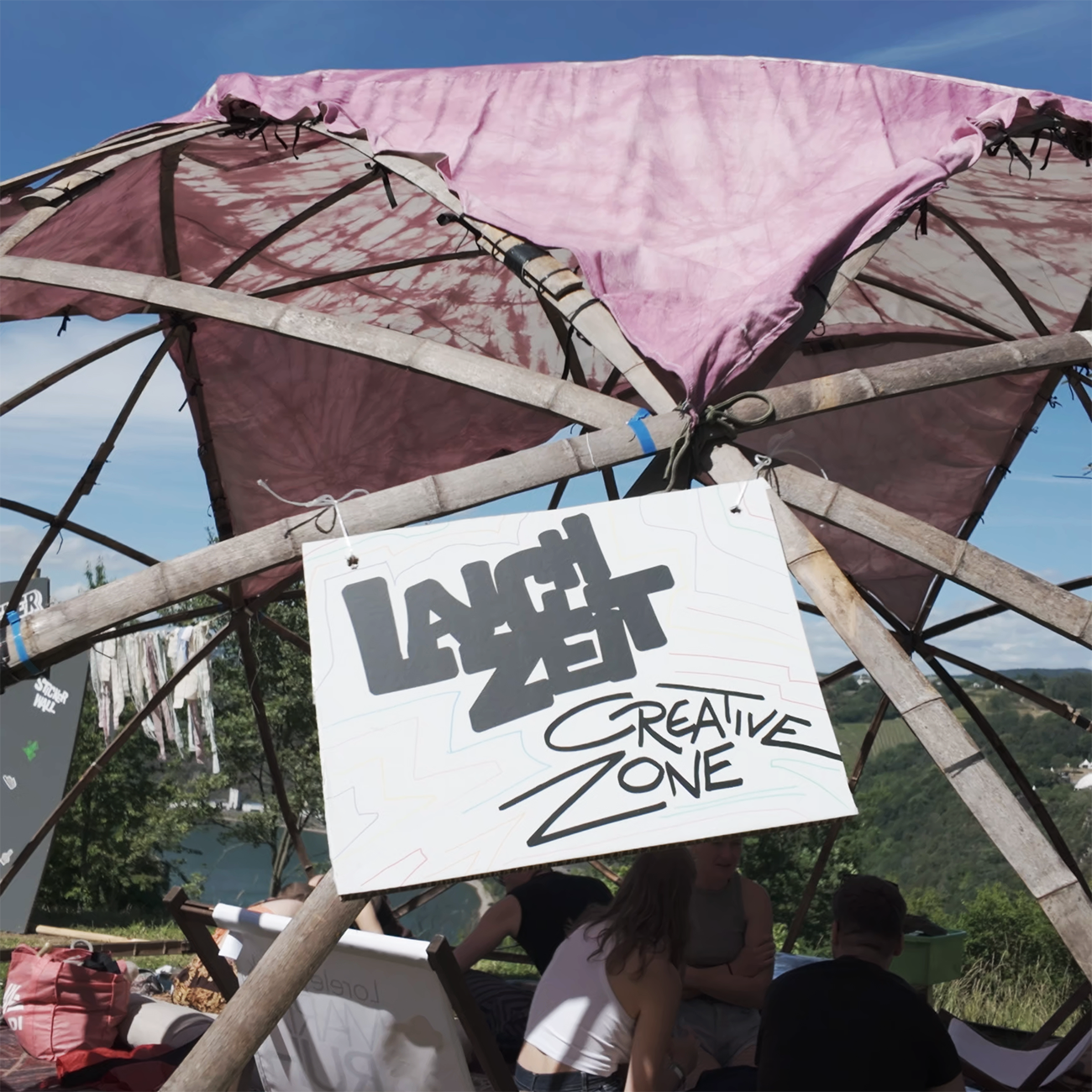 People sitting in a a tipi tent with a batik patterned red cover and a sign with the text "LANCHZEIT creative zone" with a river and trees in the background
