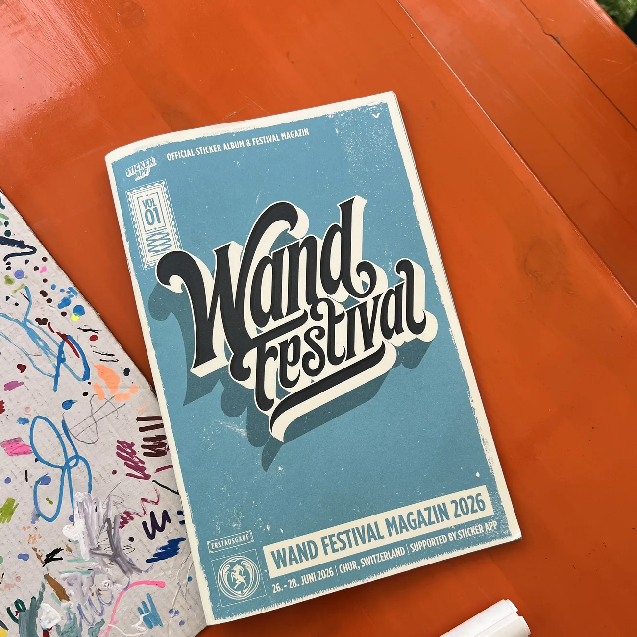 Wand Festivals offisielle klistremerkealbum og festivalmagasin 2026, støttet av StickerApp, ved siden av en malingspenn på et oransje bord