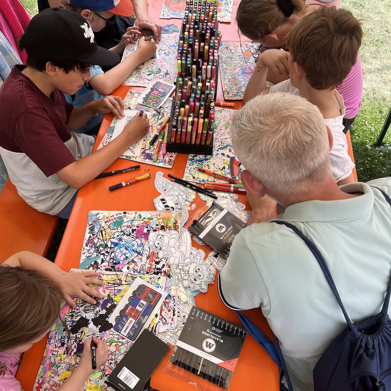Barn fargelegger die-cut klistremerker fra Brainfart rundt et stativ med malingspenner på en festivalworkshop utendørs