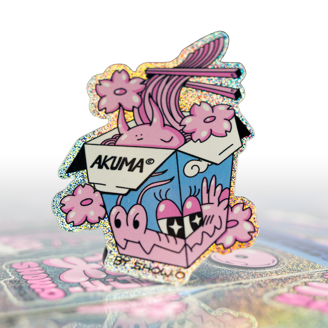 Sparkly die cut sticker with a cartoon noodle box character, pink flowers, and "AKUMA" text, set against a blurred background with similar stickers.