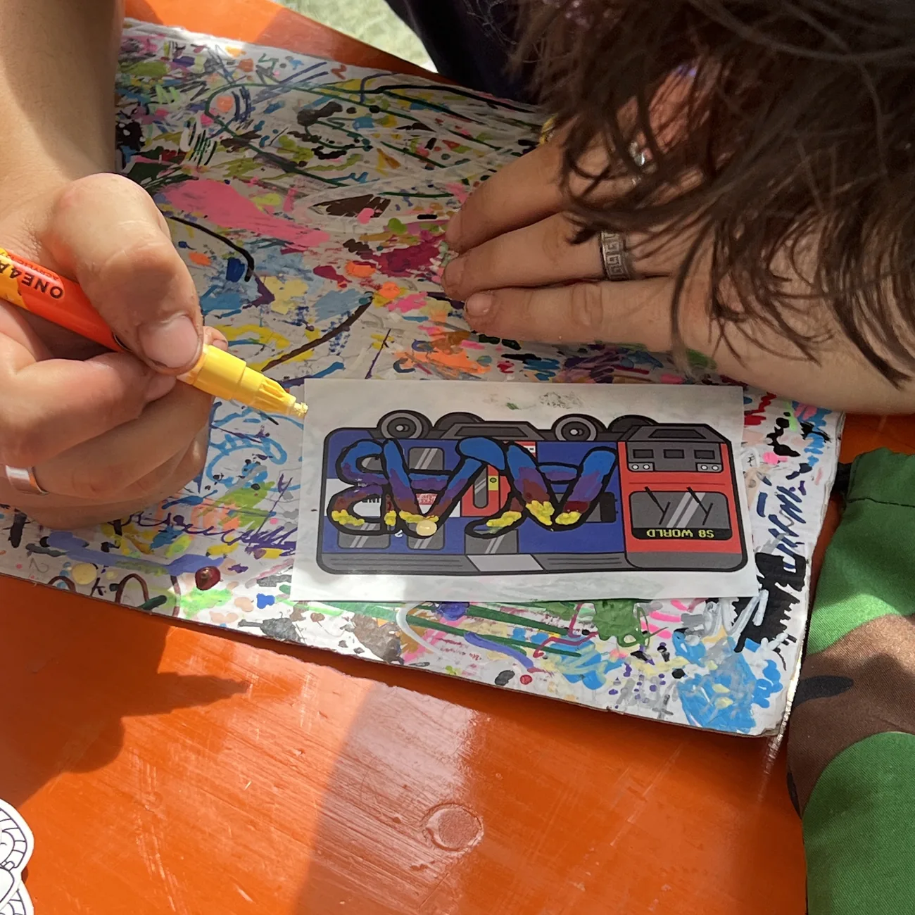 En hånd fargelegger en etikett med S8 World-toget med gul malingspenn på workshoppen til Brainfart x StickerApp