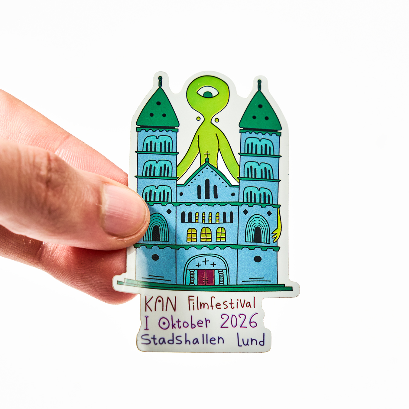 Hånd som holder et die-cut klistremerke for KAN Filmfestival, oktober 2026 i Stadshallen Lund, med en katedral og et grønt enøyd romvesen