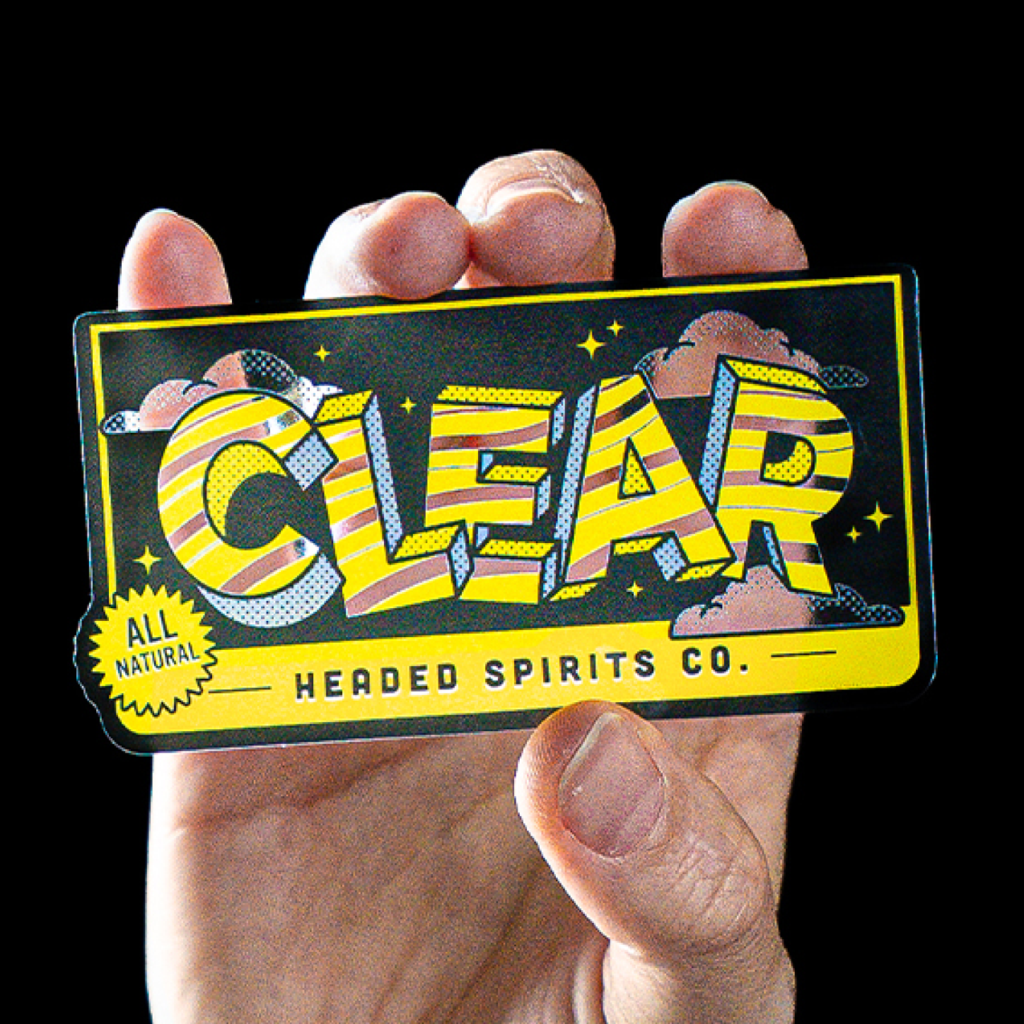 Gjennomsiktig klistremerke med gult og svart "Clear Headed Spirits Co."-design, holdt i en hånd