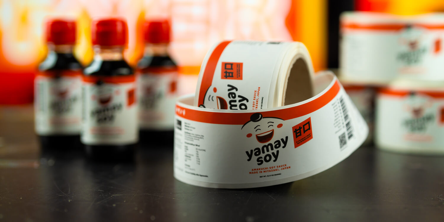 A roll of soy brand labels with vibrant artwork in the foreground, with blurred soy product bottles and additional rolls in the background.