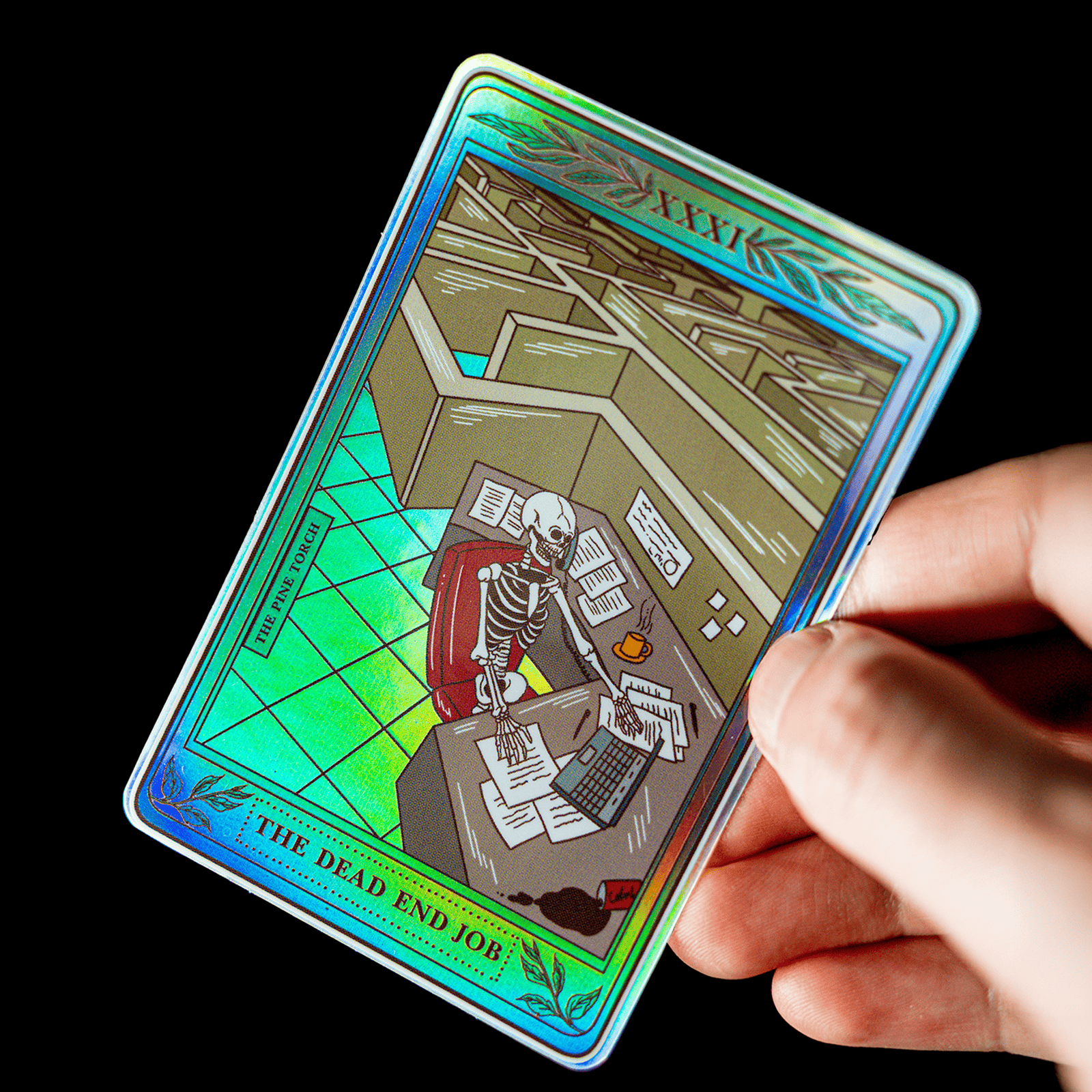 Hånd som holder et holografisk tarotkort-klistremerke med tittelen «The Dead End Job» og et skjelett ved en kontorpult