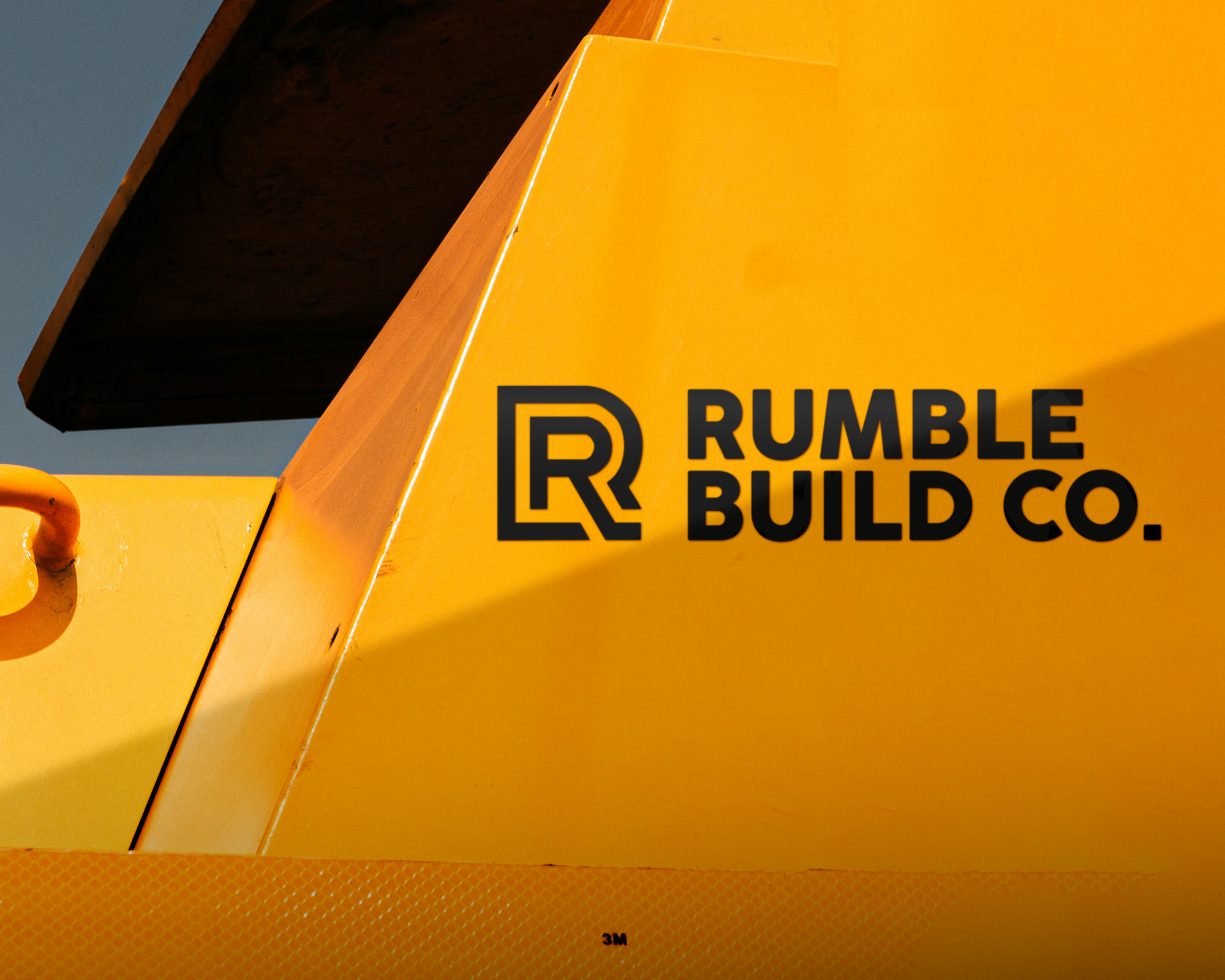 Close-up of a yellow industrial vehicle with "Rumble Build Co." vinyl transfer in bold letters on its surface, featuring an abstract "R" design.