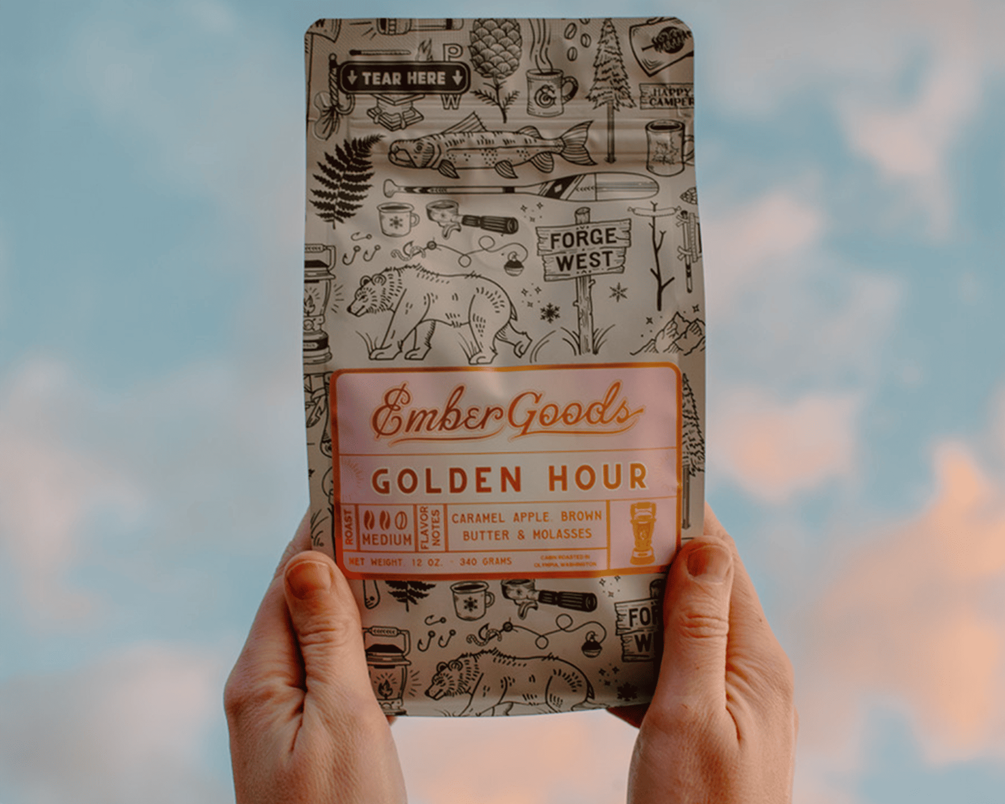 Hender som holder en pose Ember Goods Golden Hour-kaffe med metallisk etikett mot en himmel i bakgrunnen
