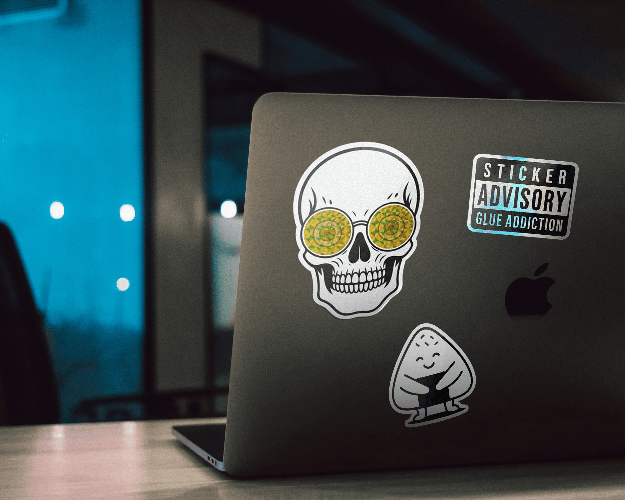 Laptop with skull, glue advisory, and rice ball stickers on the back, set on a table with a blurred blue background.