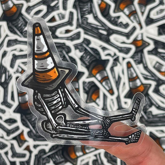 Transparent die-cut klistremerke av et skjelett som sitter på bakken med en trafikkjegle på hodet, mot en bakgrunn av klistremerker.